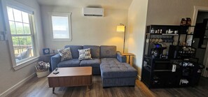 A living room with a grey sofa, a wooden coffee table, a lamp, and a kitchen area with appliances and storage shelves.