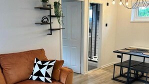 Un salon avec un canapé marron, un coussin noir et blanc, une table à manger avec de l’assiette, et une plante posée sur une étagère.