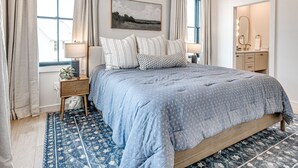 Ein Schlafzimmer mit einem großen Bett, einem Nachttisch, einem Fenster mit Vorhängen, einem Wandbild und einem Badezimmer mit Spiegel und Handtuchhalter.