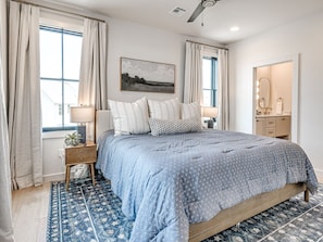 Een slaapkamer met een groot bed, nachtkastje, raam met gordijnen, een wanddecoratie en een badkamer met spiegel en handdoekhouder.