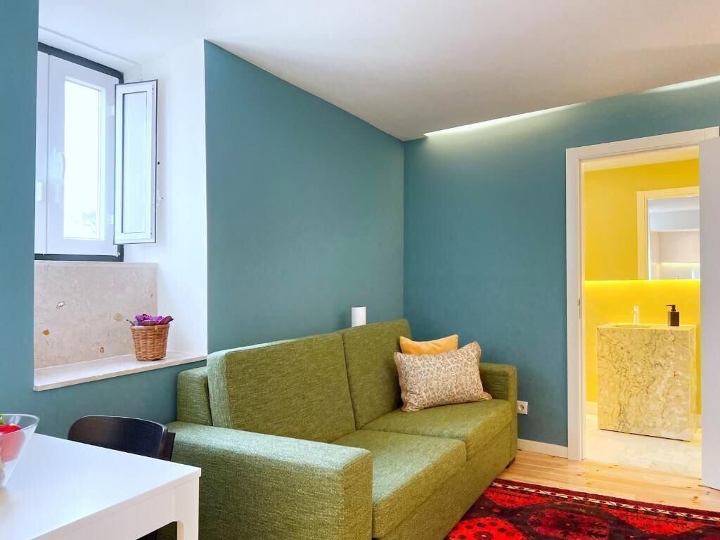 A living room with a green sofa, a small table with a bowl, a window with a basket, and a yellow bathroom in the background.