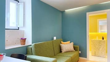 A living room with a green sofa, a small table with a bowl, a window with a basket, and a yellow bathroom in the background.