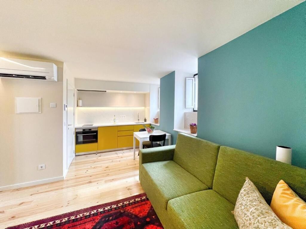 A modern apartment with a green sofa, a kitchen with yellow cabinets, and a red patterned rug.