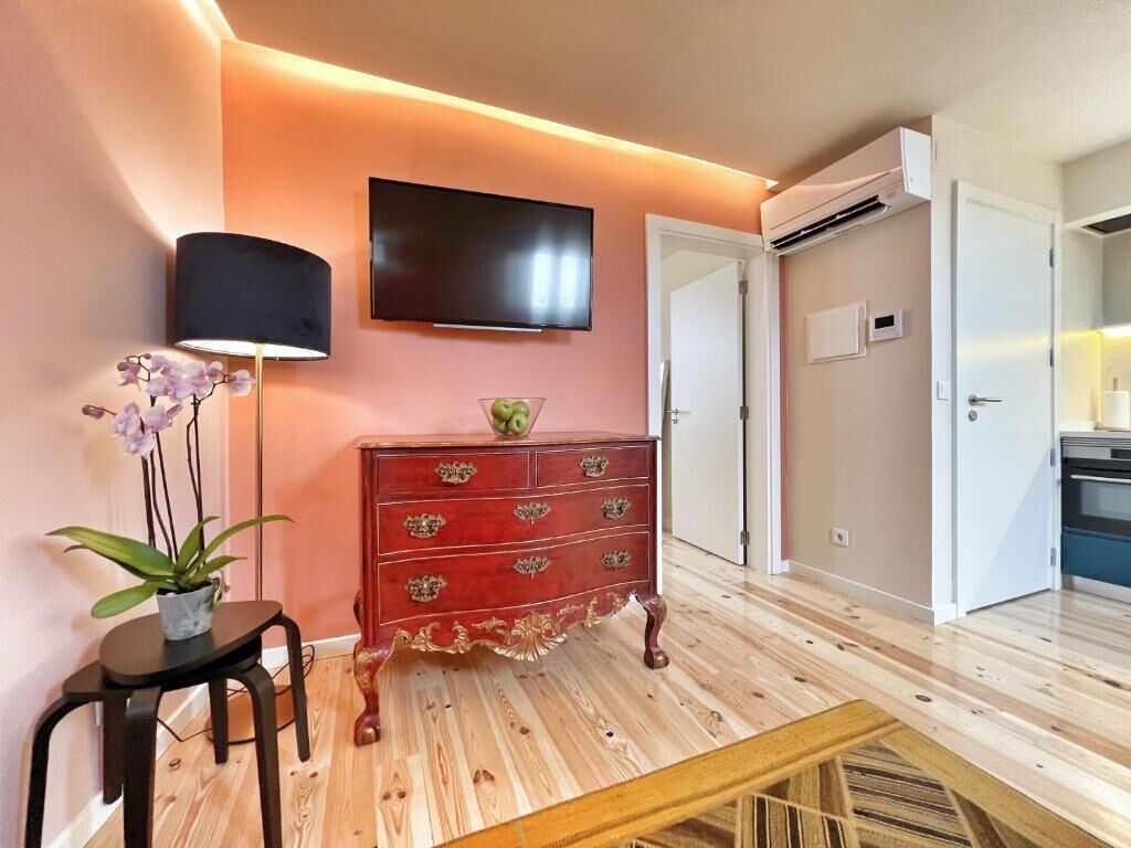 A room with a wooden floor, a red dresser with ornate handles, a flat-screen TV mounted on the wall, a potted plant, and a small table.