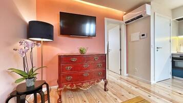 A room with a wooden floor, a red dresser with ornate handles, a flat-screen TV mounted on the wall, a potted plant, and a small table.