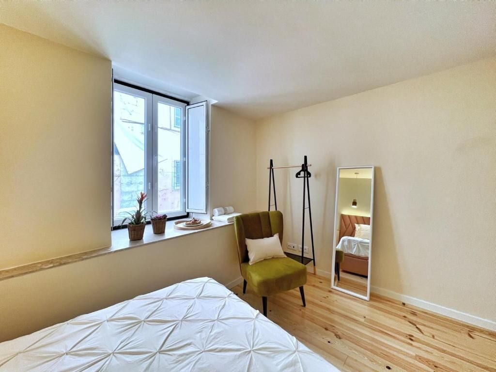 A bedroom with a bed, a chair, a window, a mirror, and a small table with a plant.