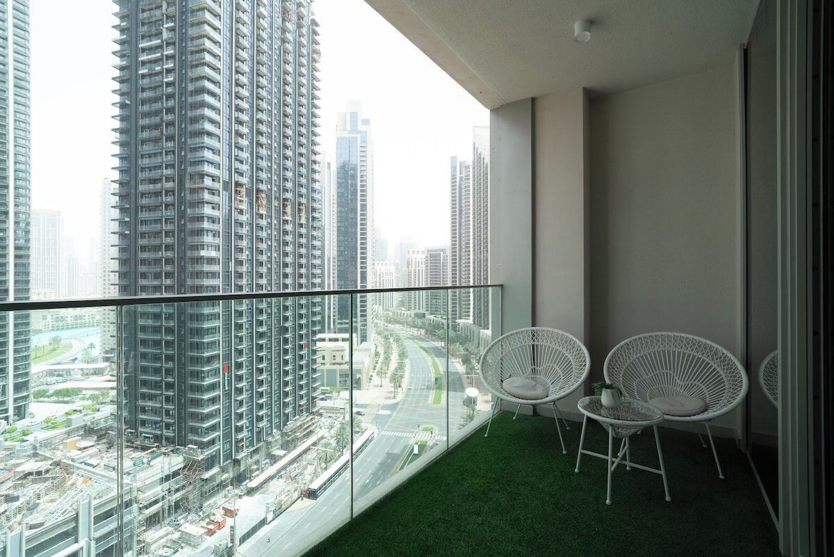 Uma varanda com vista para edifícios altos, um tapete verde e cadeiras brancas.
