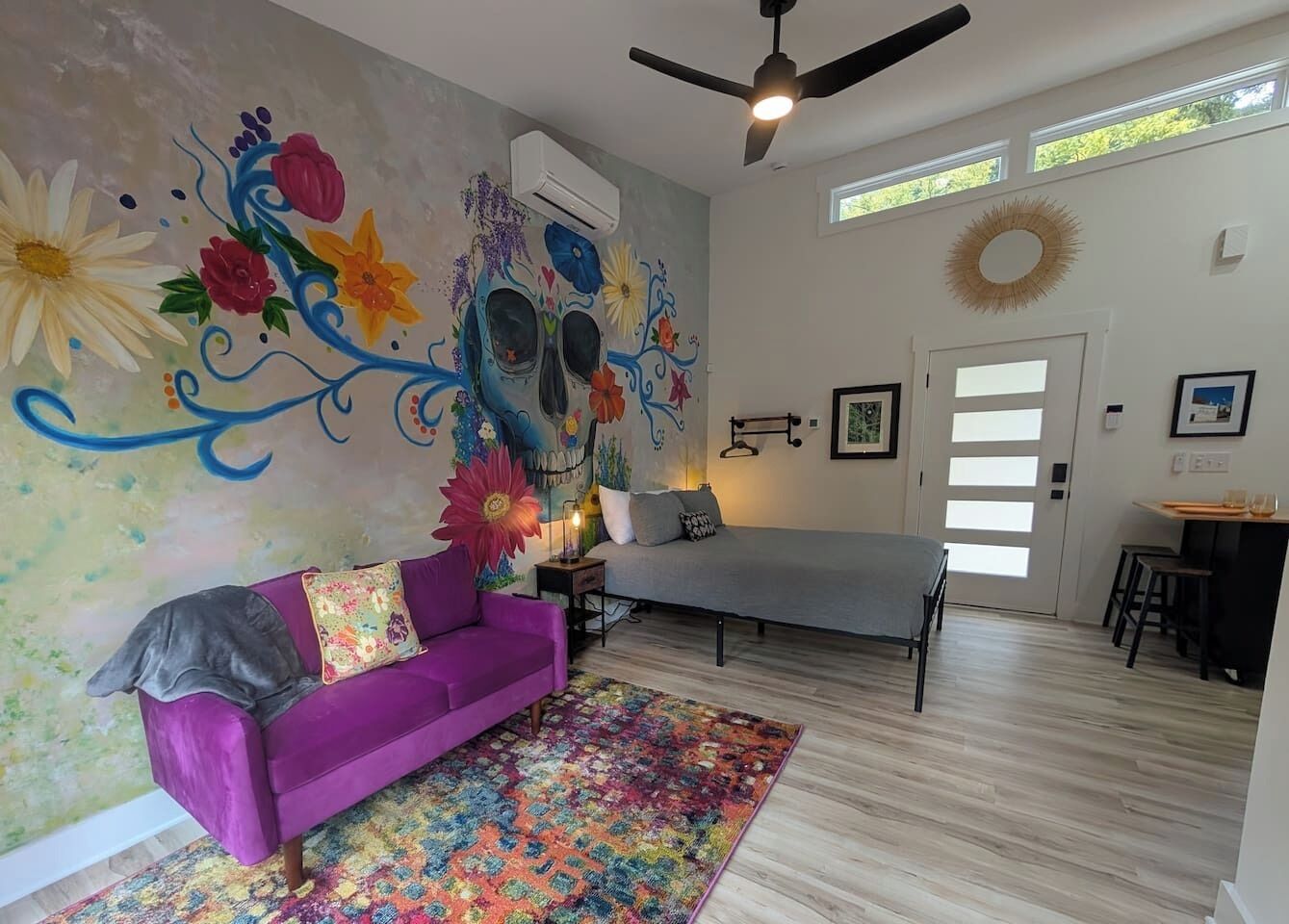 A room with a purple sofa, a bed, a colorful rug, and a wall mural.