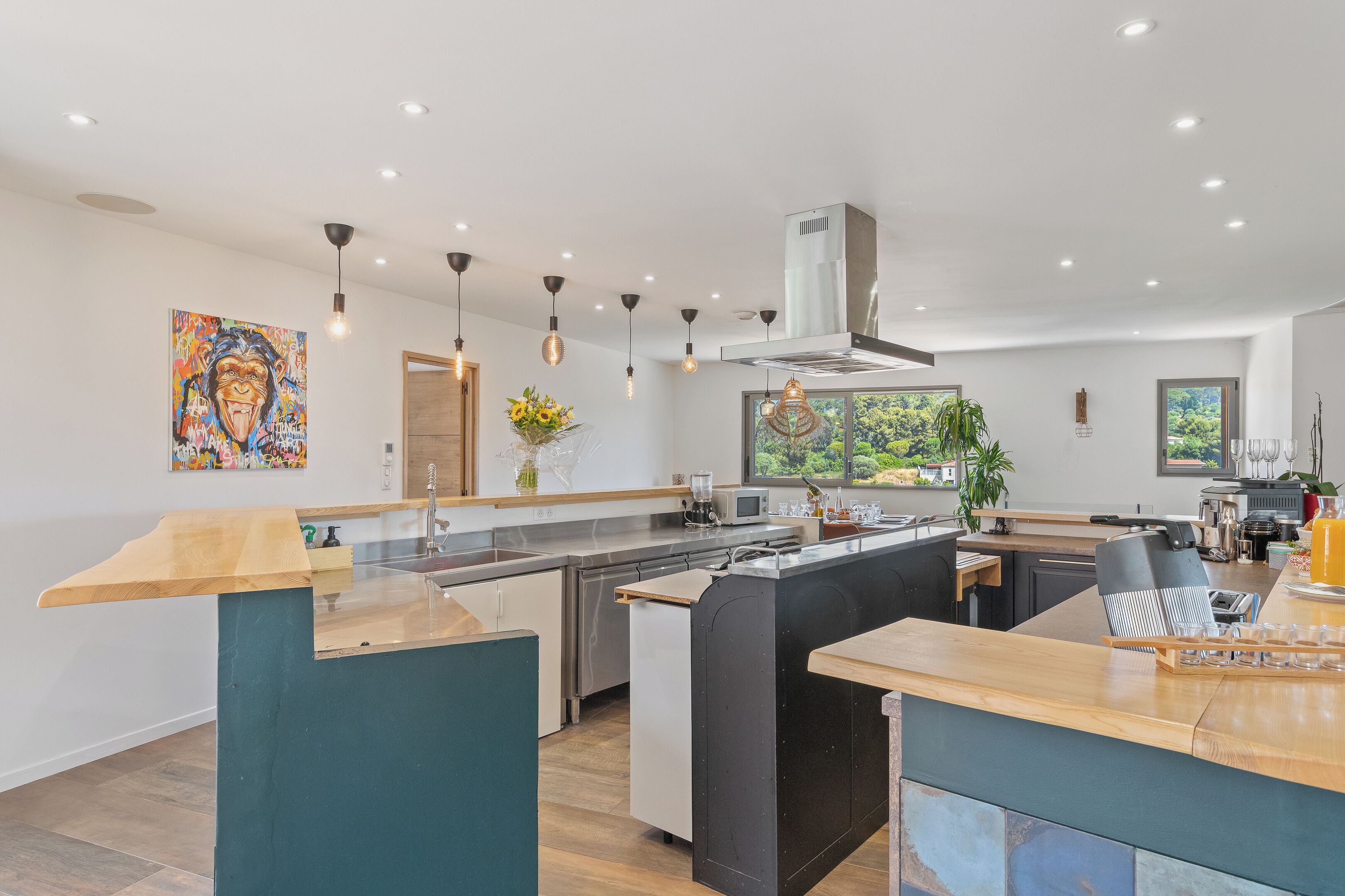 A modern kitchen with a central island, pendant lighting, and a large window offering a view of greenery.
