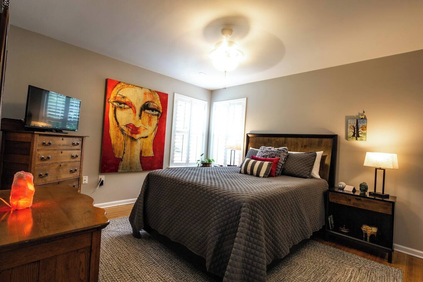 A bedroom with a bed, a TV, a painting, and a nightstand with a lamp.