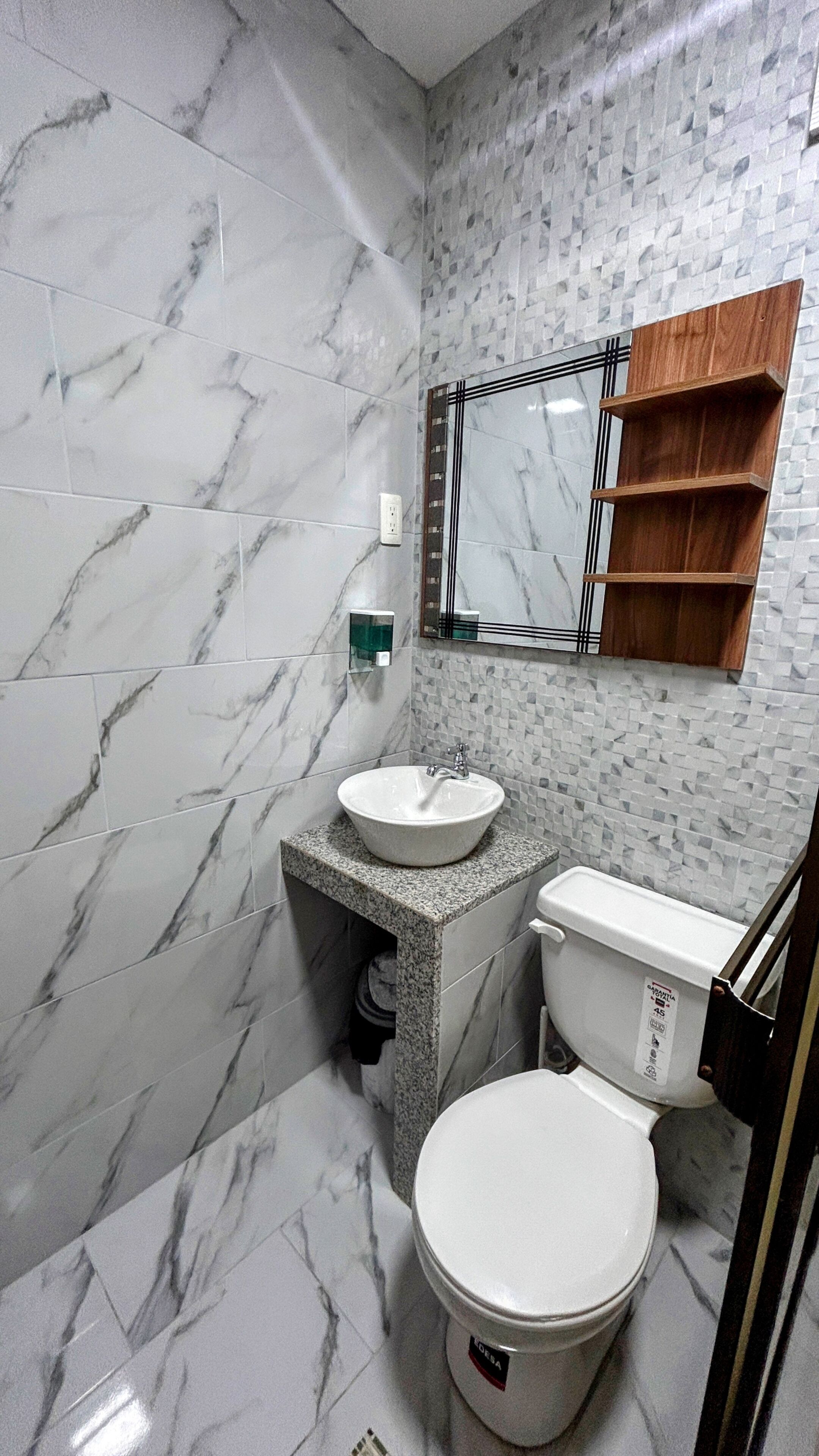 Ein Badezimmer mit Marmorboden, einem weißen Waschbecken, einer Toilette und einem Spiegel mit einem Holzregal darüber.