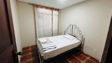 Ein kleines Zimmer mit einem Einzelbett, einem Metall-Kopfteil, einem Fenster mit transparenten Vorhängen und einem gemusterten Boden.