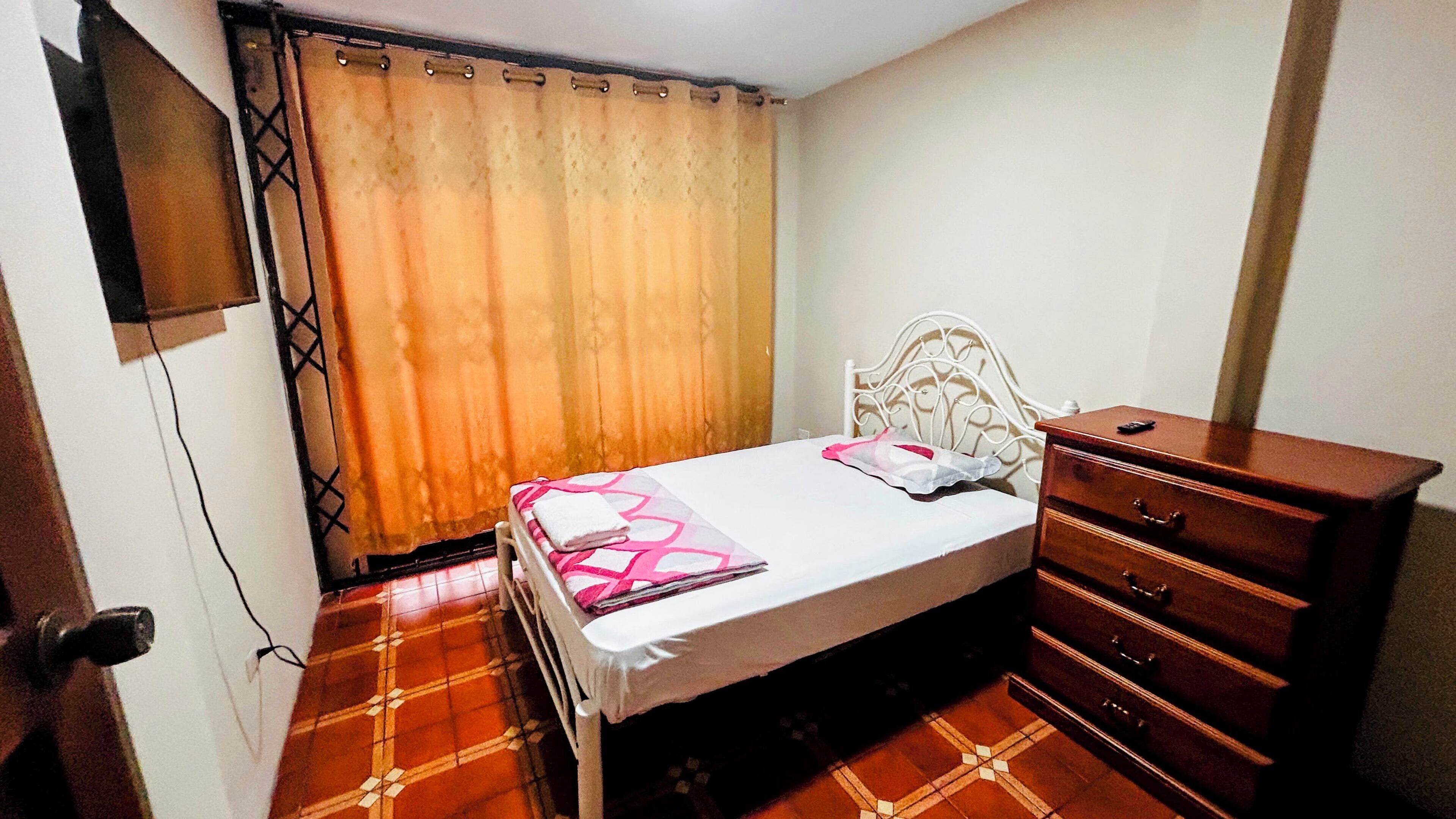 Quarto com cama, cômoda, televisão na parede e cortina.