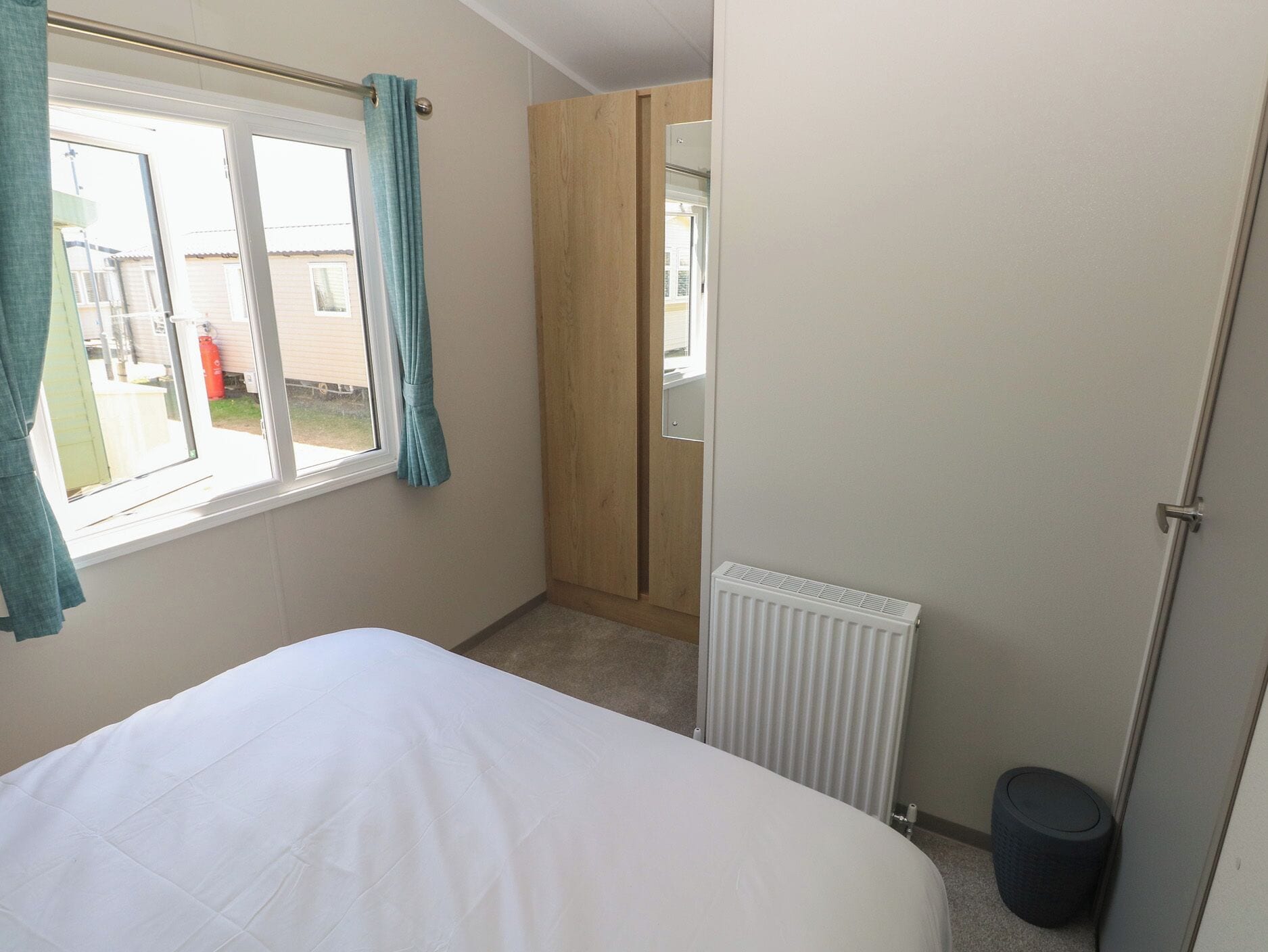 A bedroom with a bed, a window with curtains, a radiator, and a door leading to a hallway.