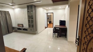 Uma sala de estar ampla com piso de mármore, uma estante embutida, uma mesa de jantar e uma porta que dá para outro cômodo.