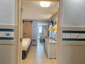 Un couloir étroit mène à une chambre qui contient un lit, une commode et une petite table avec une lampe.