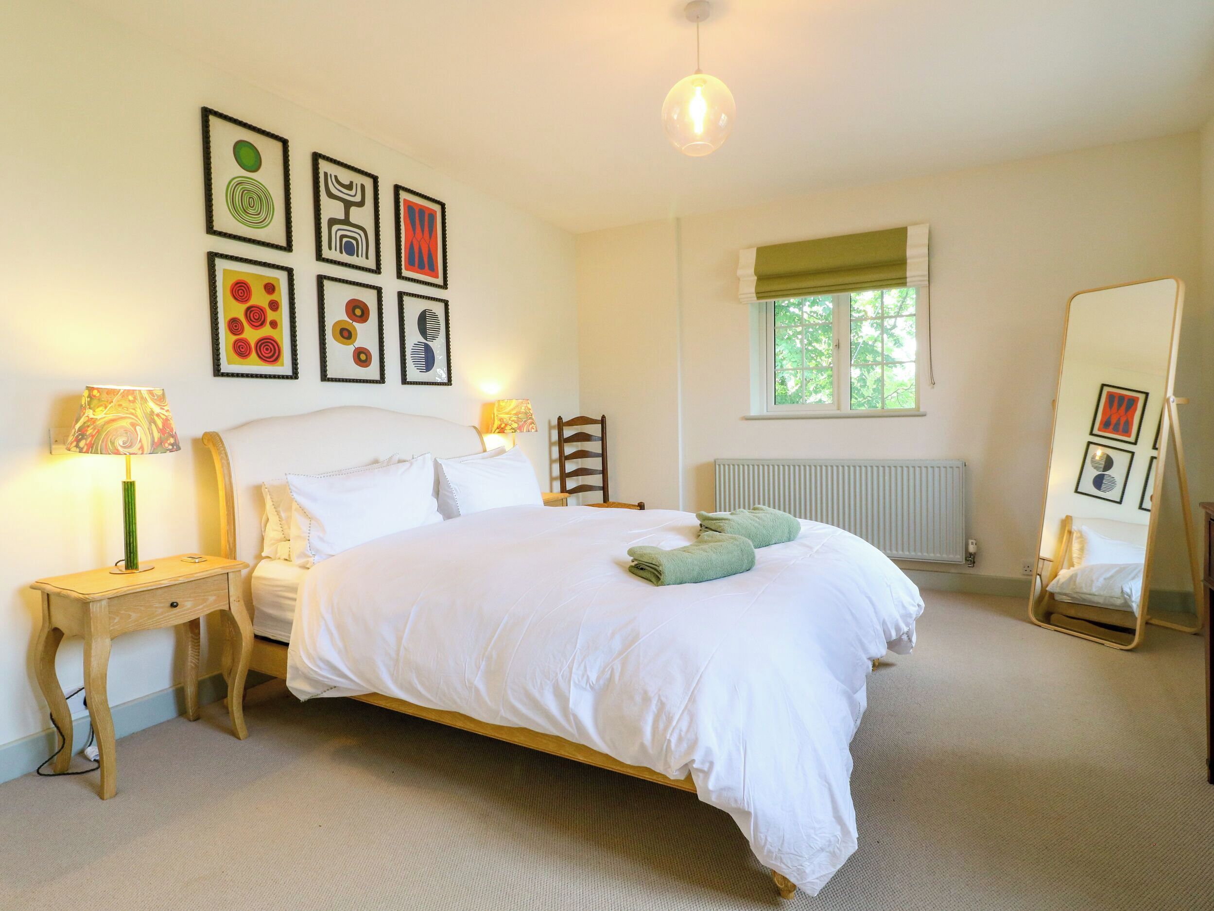 A bedroom with a bed, bedside tables, a mirror, a window, and wall art.