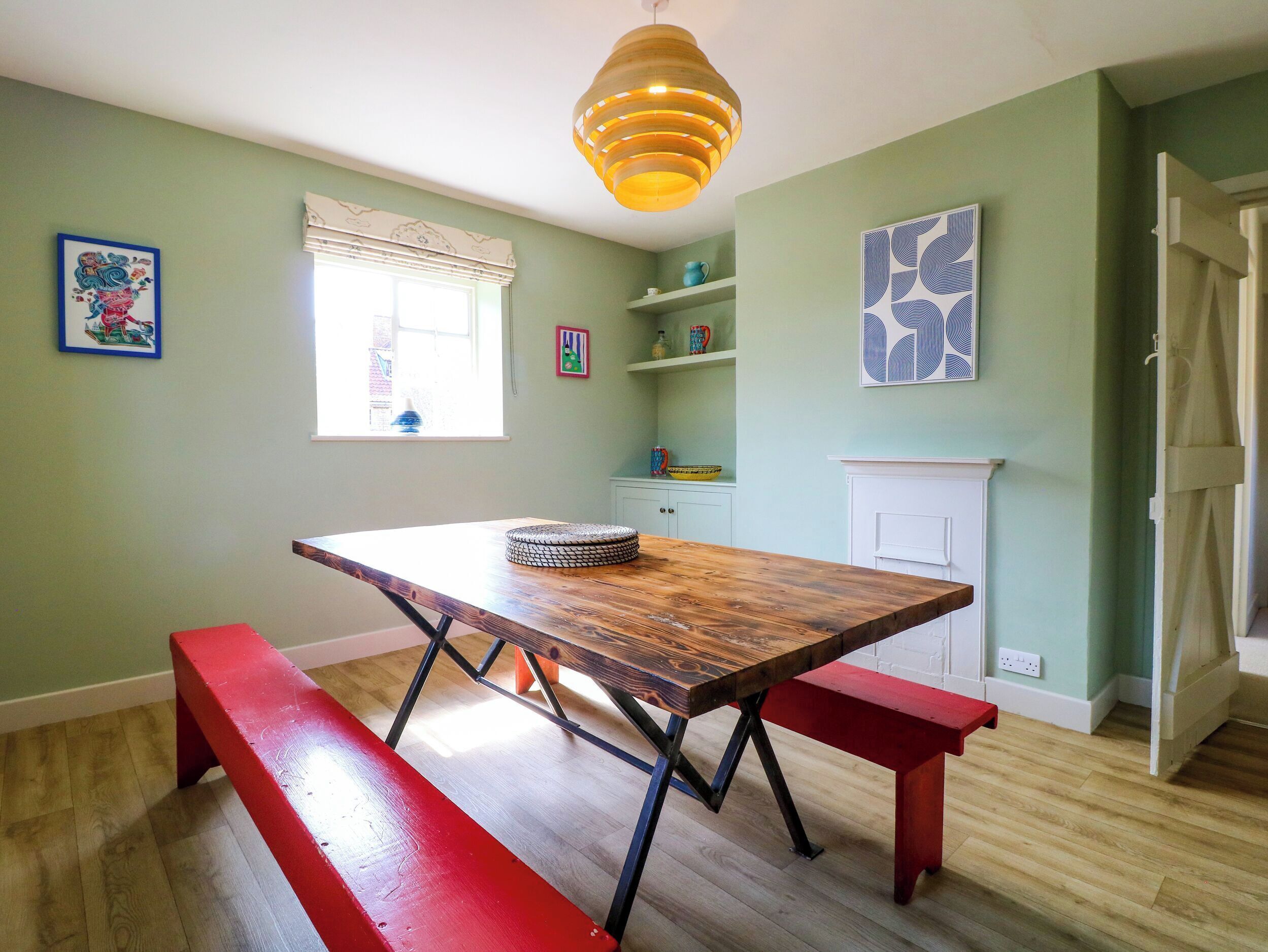 A dining area with a wooden table, benches, a hanging light fixture, a window with blinds, and a fireplace.