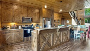 A rustic kitchen with wooden cabinets, a central island, stainless steel appliances, and a dining area with chairs. - Sweet Sunrise | Luxury Cabin w/ Slide, Hot Tub & Views (Broken Bow)