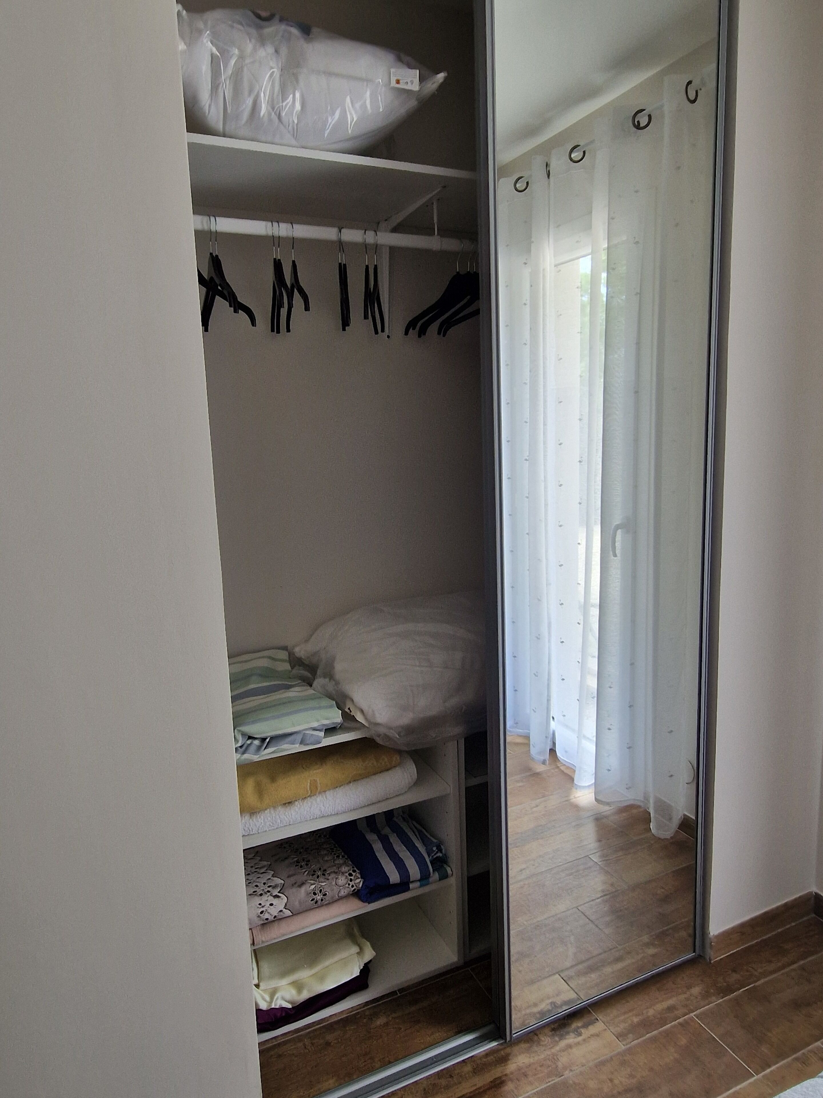 A bedroom with a wardrobe, a bed with pillows, a mirror, and a window with curtains.