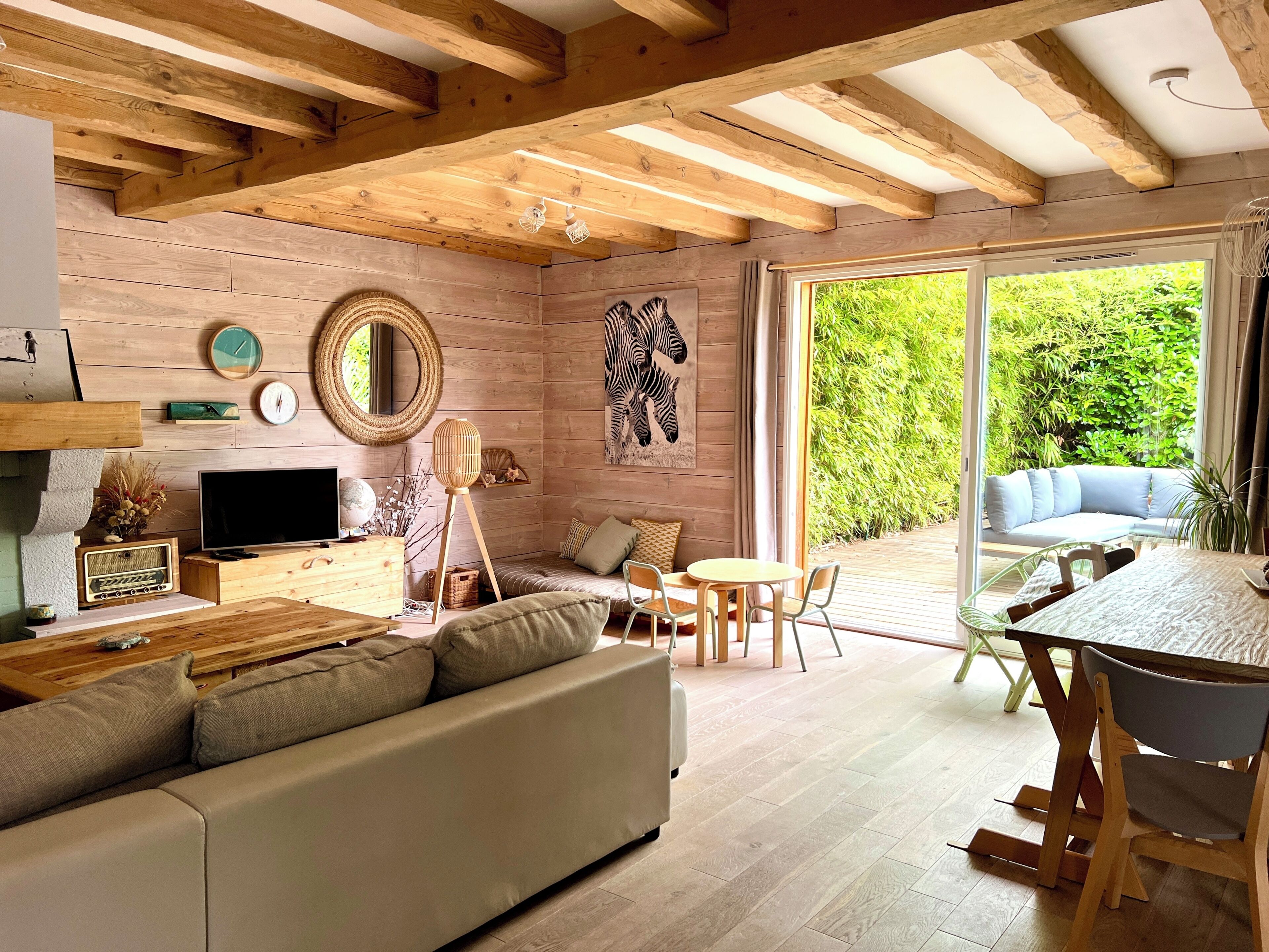 Un salon chaleureux avec un canapé, une télévision, des poutres en bois, un coin repas avec une table et des chaises, et une grande fenêtre donnant sur un paysage verdoyant.
