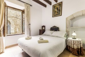 A bedroom with a large bed, a nightstand, a lamp, a painting, and a stone wall.