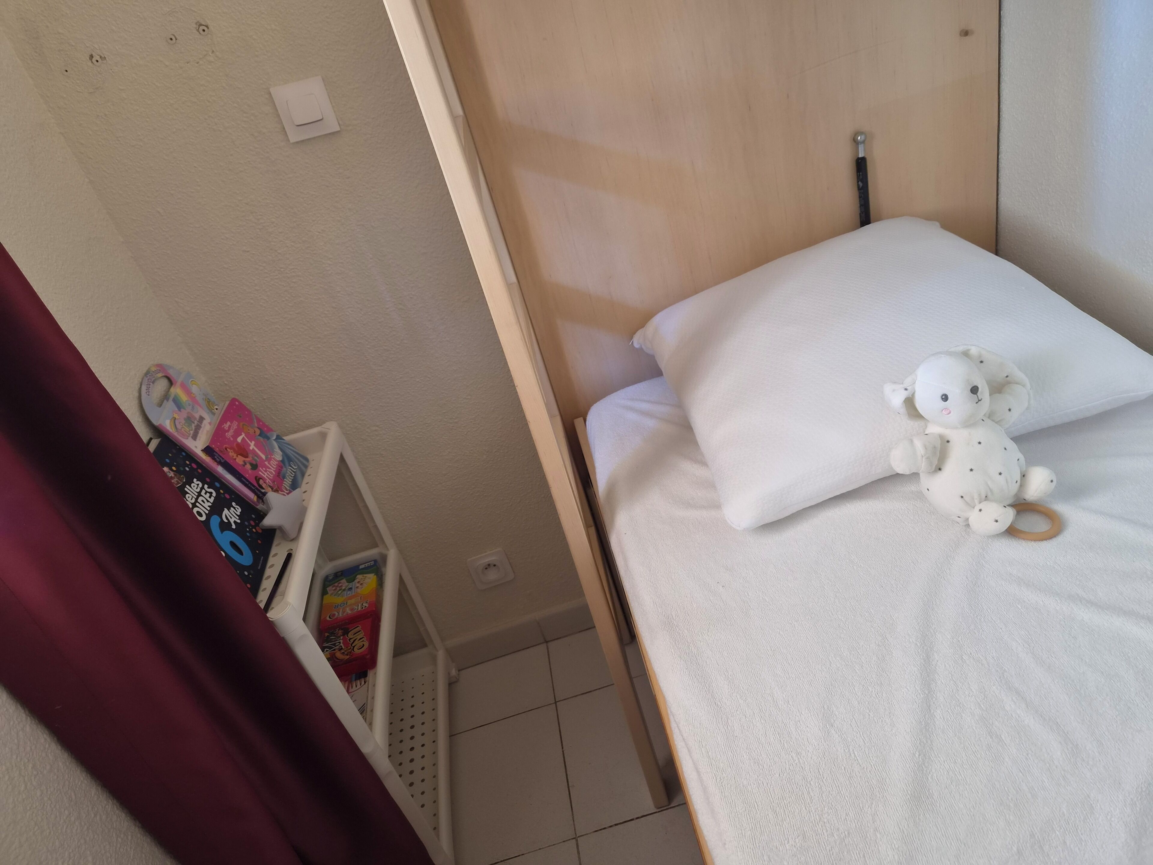 Un lit avec un oreiller blanc et un jouet en peluche, une étagère avec des livres et un petit panier, et un mur avec un interrupteur et une prise de courant.
