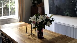 Ett matrum med ett träbord, stolar, en kristallkrona, ett fönster med gardiner, en krukväxt och en stor tavla på väggen.