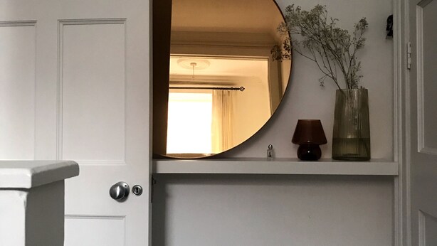 En vit dörr med en rund spegel ovanför, ett litet bord med en vas och en lampa på väggen.