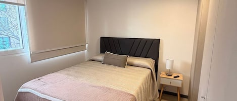 Un dormitorio con cama, mesita de noche, lámpara y ventana.