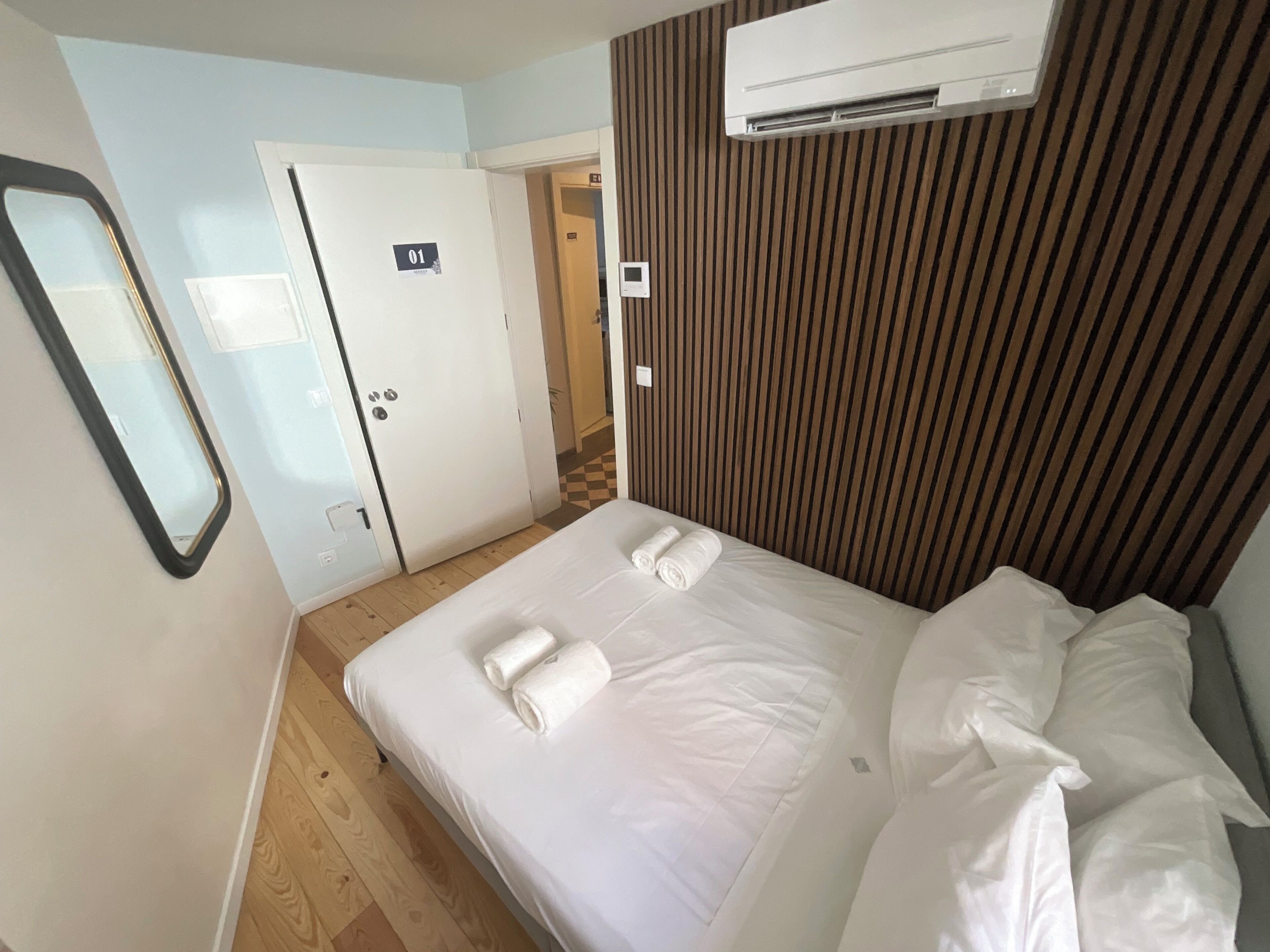 A hotel room with a bed, a mirror, an air conditioning unit, and a door.