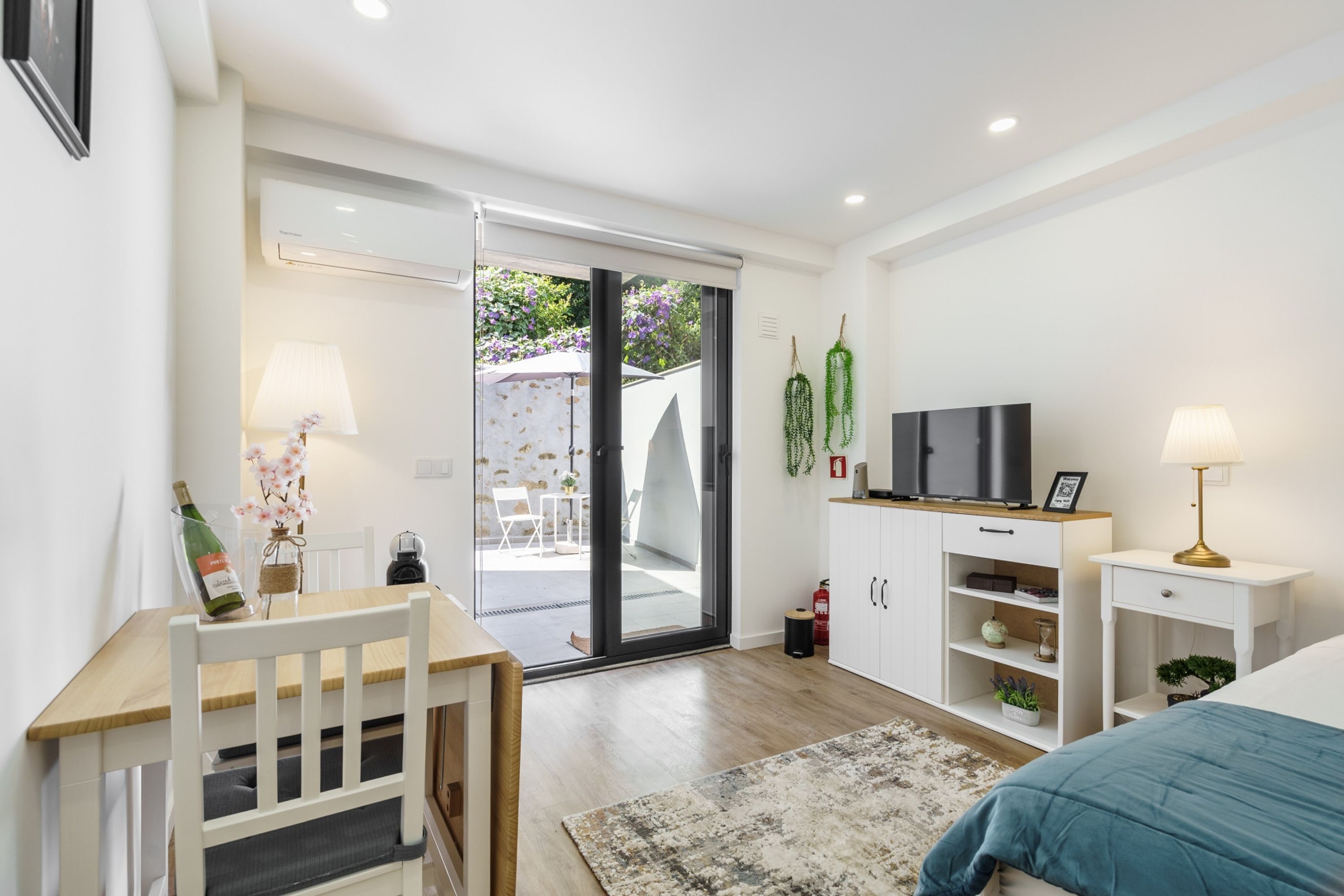Un dormitorio moderno con cama, televisor, escritorio con silla, puerta corrediza de vidrio que da a un balcón y un mueble bajo.