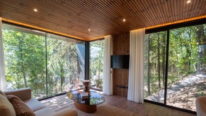 A modern living room with a round coffee table, a sofa, and a TV mounted on the wall. Large windows offer a view of a forested area. - Prendal Eco Living Villa IV by Minho s Guest (Fafe)