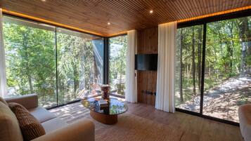 A modern living room with a glass wall, a round coffee table, and a flat-screen TV.