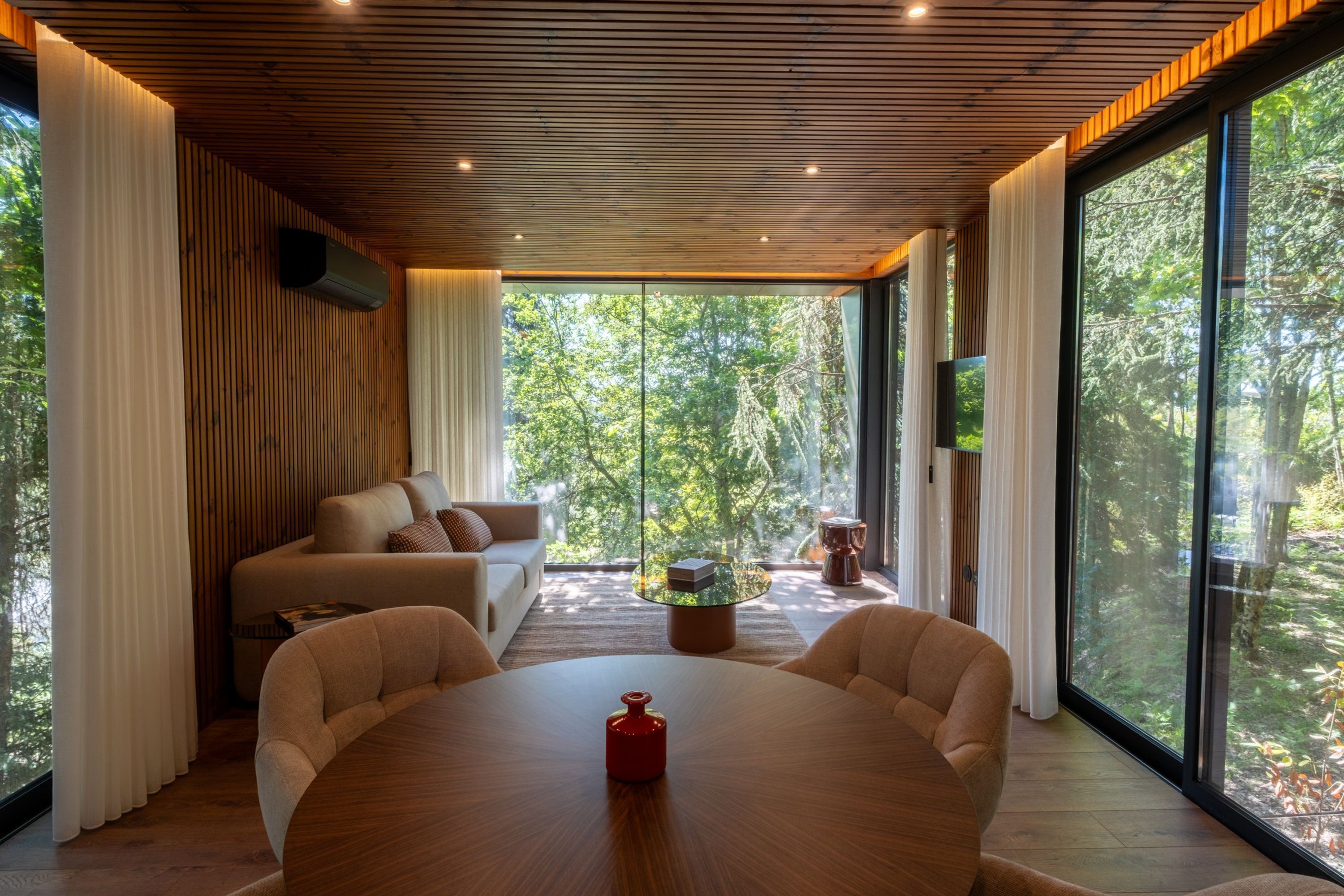 A modern interior with a wooden ceiling, large windows, a round table, and a sofa.