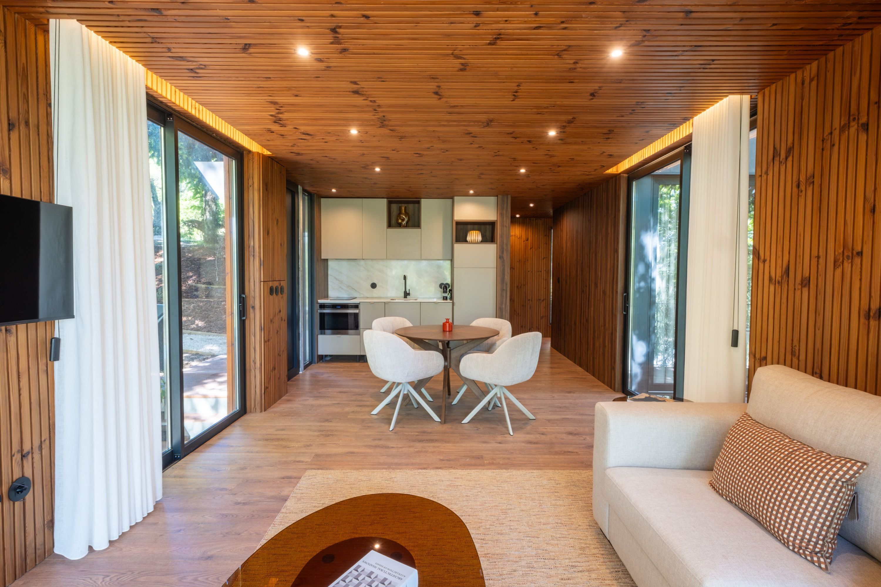 A modern interior with a wooden ceiling, a dining area with white chairs, a sofa with a patterned pillow, and a kitchen in the background.