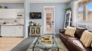 Una sala de estar moderna con un sofá de cuero marrón, una mesa de centro de vidrio y una zona de cocina con armarios blancos y un televisor.