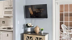 Un televisor de pantalla plana montado en una pared sobre un mueble bajo de madera con objetos decorativos, en una habitación con muebles blancos y un área de comedor visible a través de un vidrio.