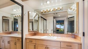 A bathroom with two sinks, wooden cabinets, and a large mirror with lights above. - Waikoloa Fairways #a101: Golf Course Ocean Views! (Waikoloa)