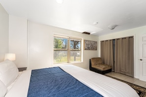 Ein Hotelzimmer mit Bett, Stuhl, Fenster mit Blick auf Bäume, Tür und einem Wandbild.
