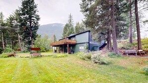 A house with a wooden deck, a staircase leading to a second floor, and a view of a forested area. - Glacier Gateway Lodge | Hot Tub & Firepit (Hungry Horse)