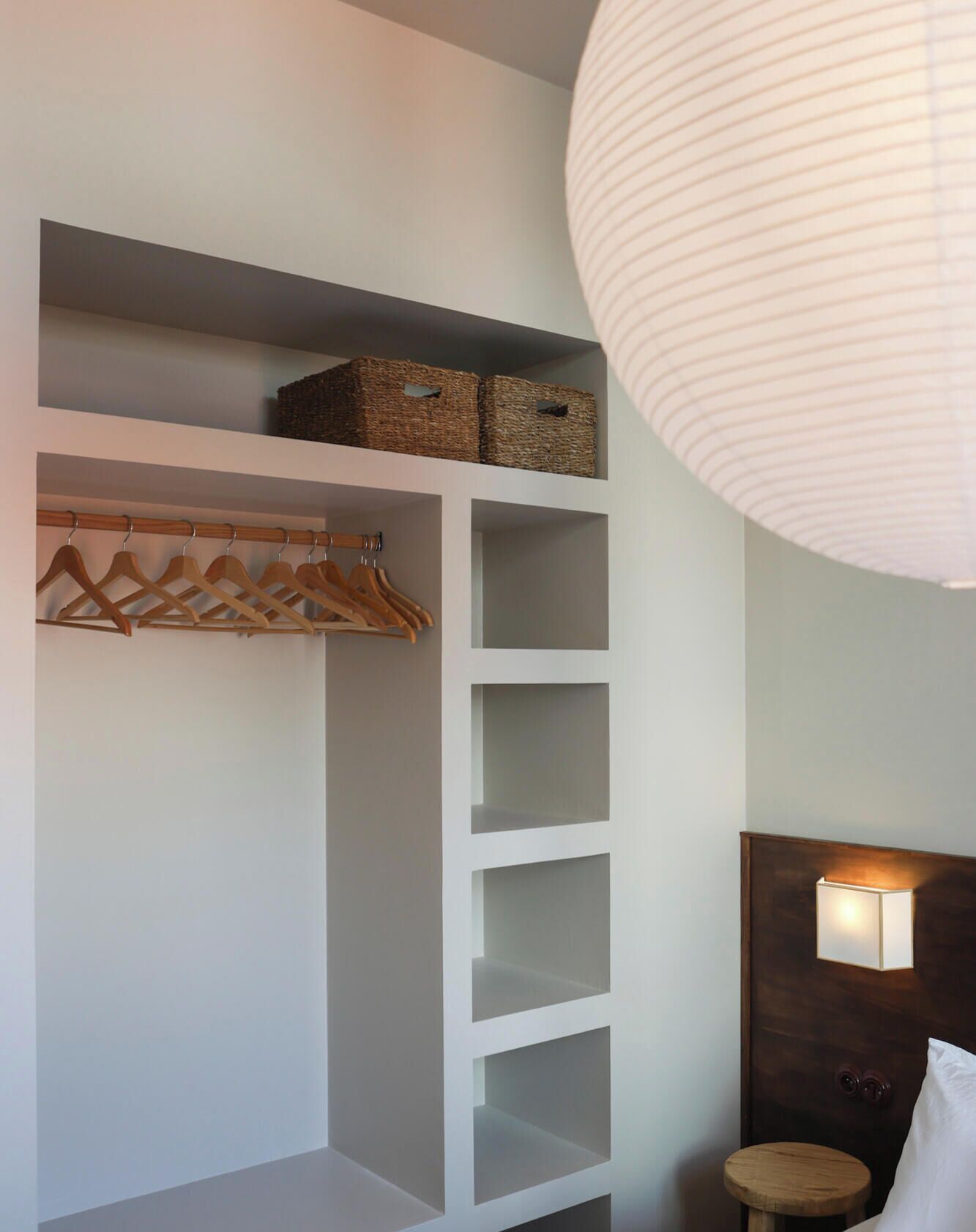 Um armário com cabides de madeira e cestos de palha, uma mesinha de cabeceira com luminária, e um lanterna de papel branca.