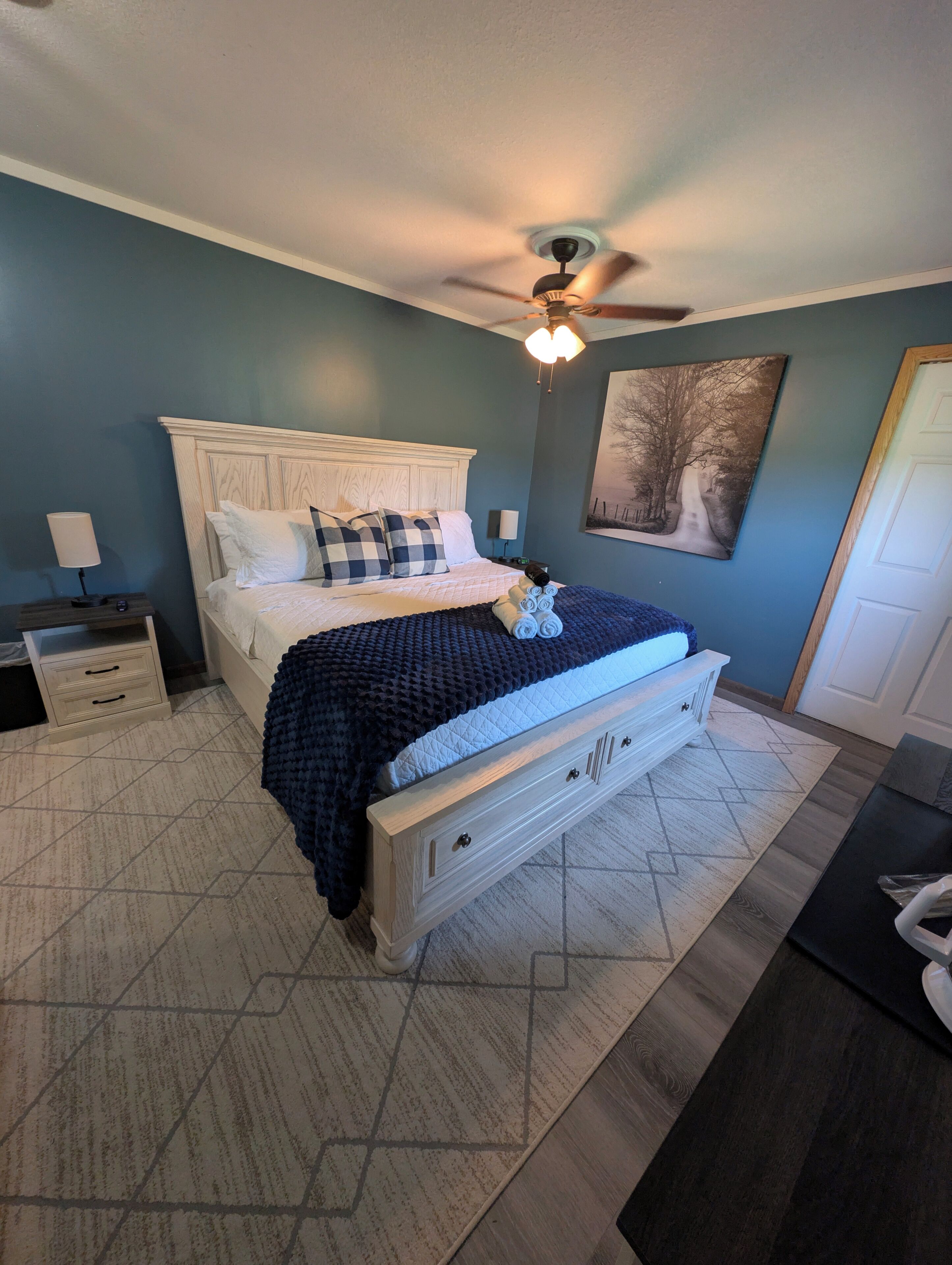 A bedroom with a bed, bedside tables, a ceiling fan, and a painting on the wall.