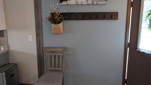 A room with a wooden chair, a wall-mounted shelf with flowers, a coat rack, and a painting of a landscape.