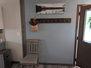 A room with a wooden chair, a wall-mounted shelf with flowers, a coat rack, and a painting of a landscape.