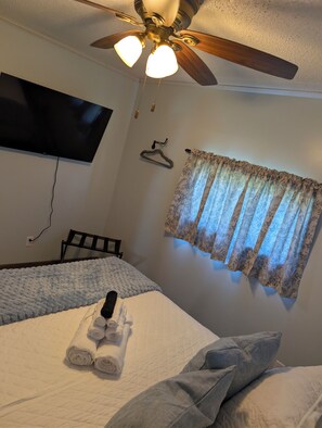 A hotel room with a bed, a ceiling fan, a TV mounted on the wall, and a window with patterned curtains.