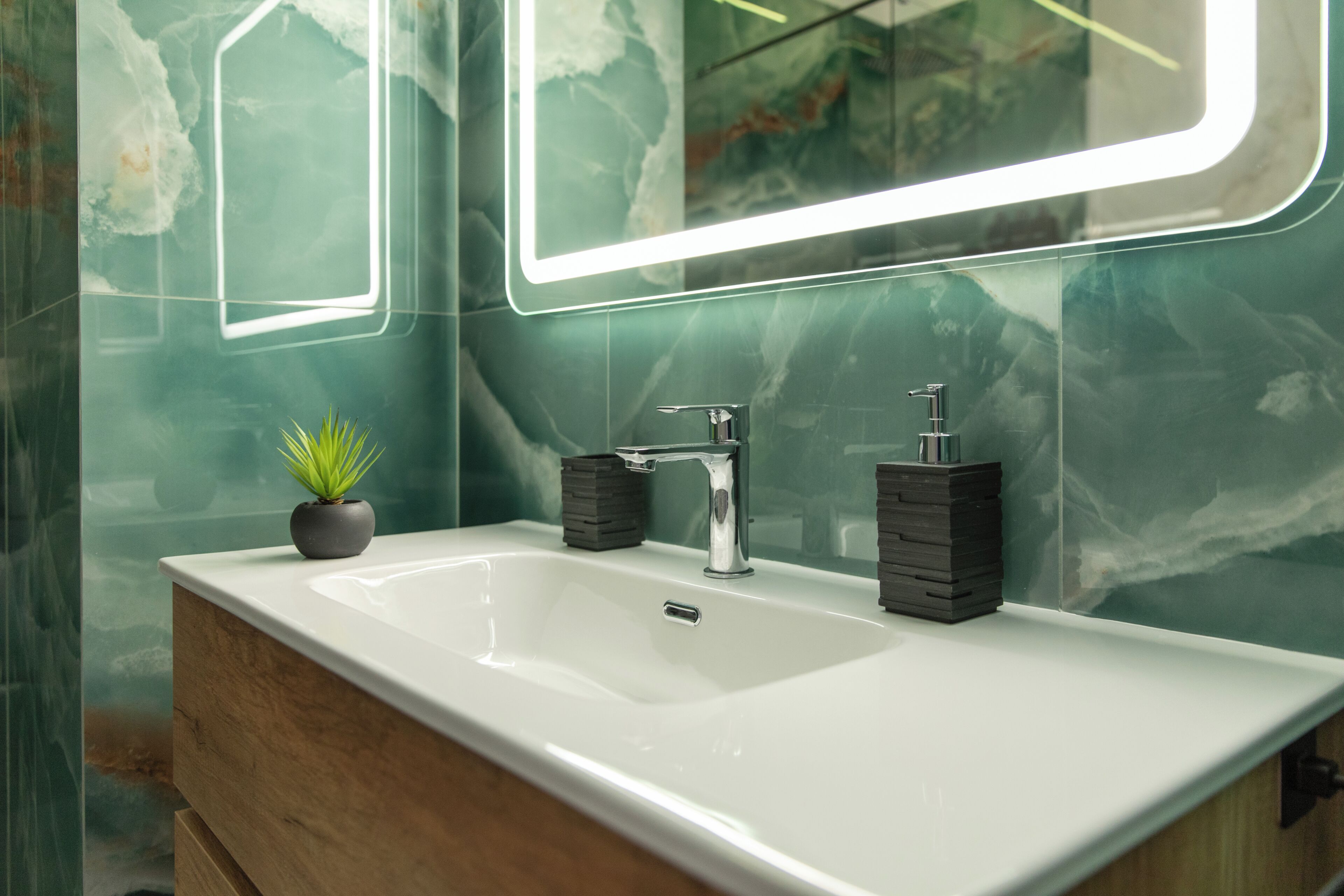 Un baño con un lavamanos blanco, dos dispensadores de jabón negros, una pequeña planta en maceta y un espejo con luz de neón.