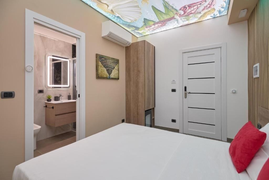 A bedroom with a bed, a bedside table, a mirror, a door, and a ceiling with a light fixture.