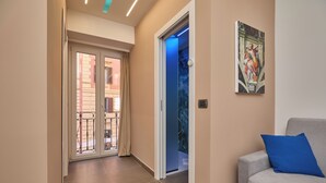 A hallway with a sliding glass door leading to a balcony, a blue-lit elevator, and a grey sofa with a blue pillow.