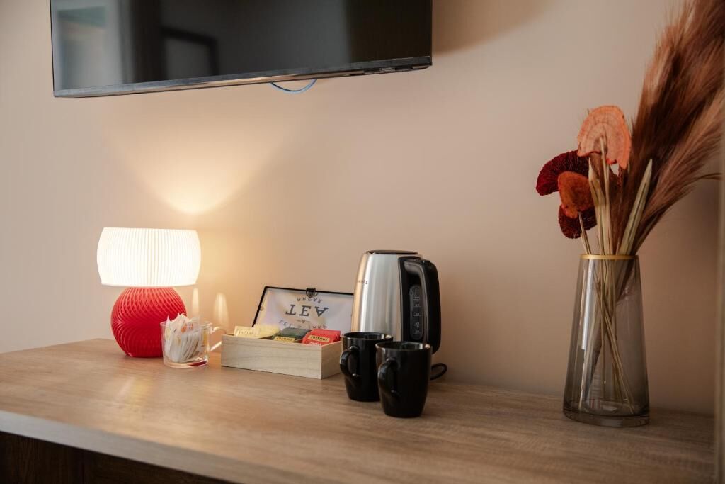 A flat-screen TV mounted on the wall, a red lamp, a toaster, two black mugs, a glass vase with dried flowers, and a framed picture on a wooden surface.